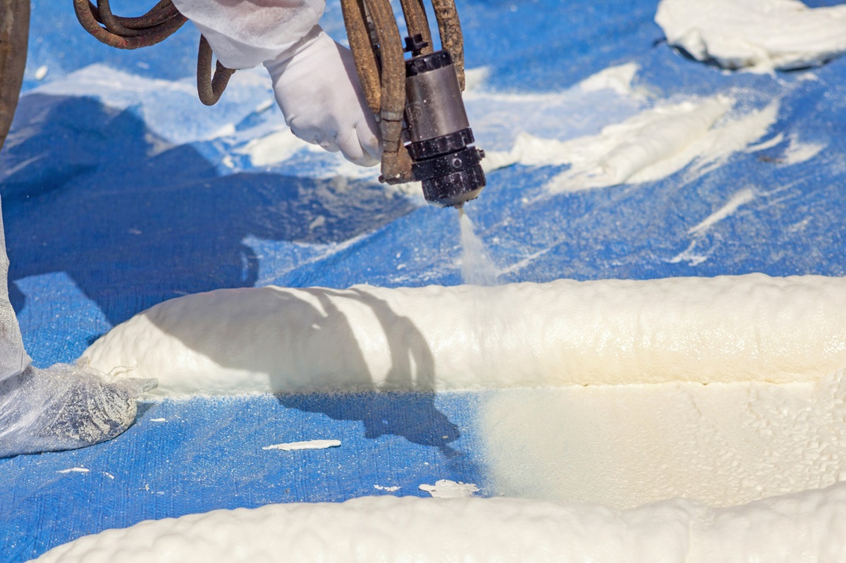 Brazos thermal worker with white gloves on coating the roof with high grade spray foam using spray gun tools.