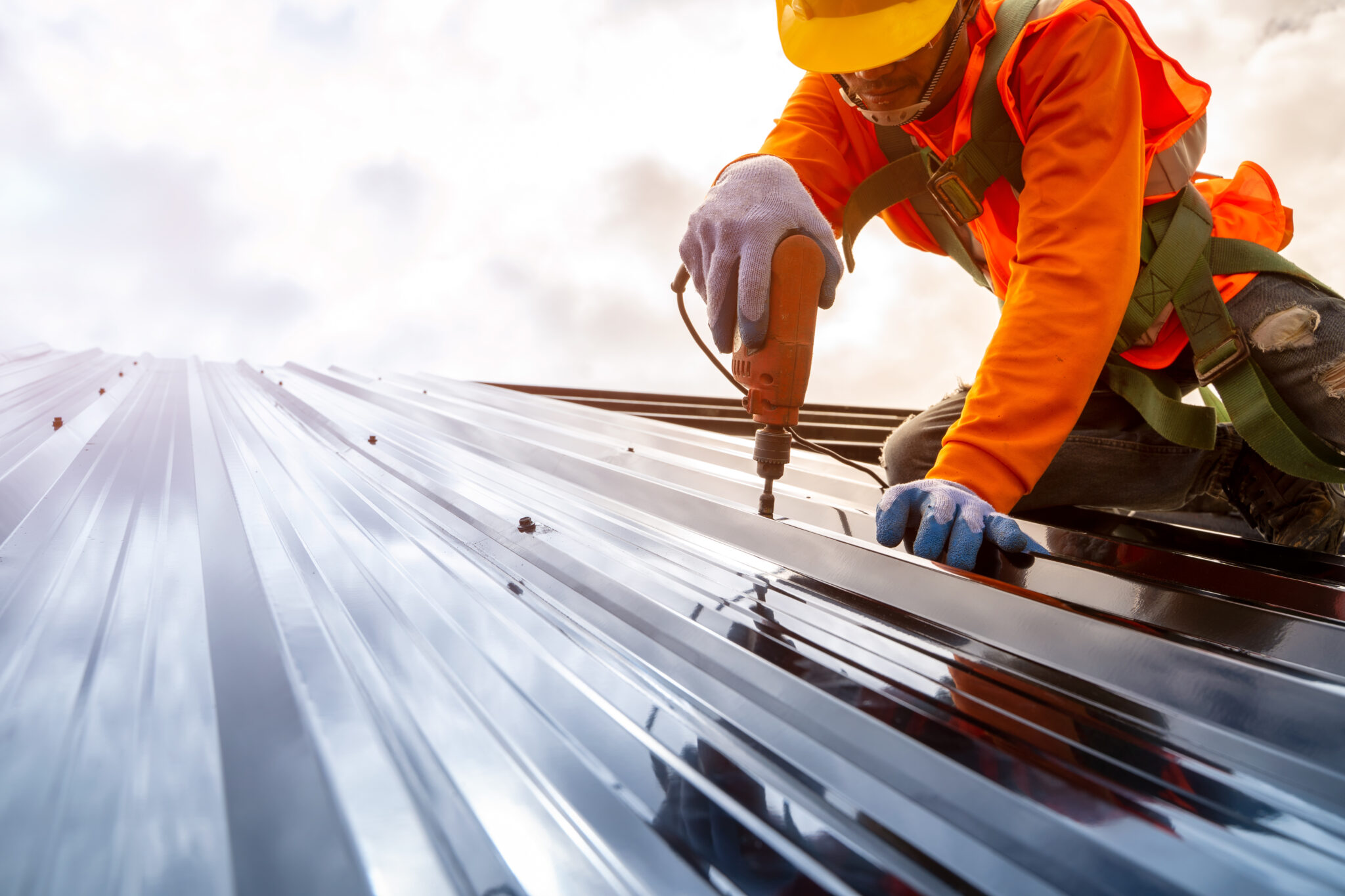 Worker on metal roof in safety gear using tool to install bolts and screws to the roof.