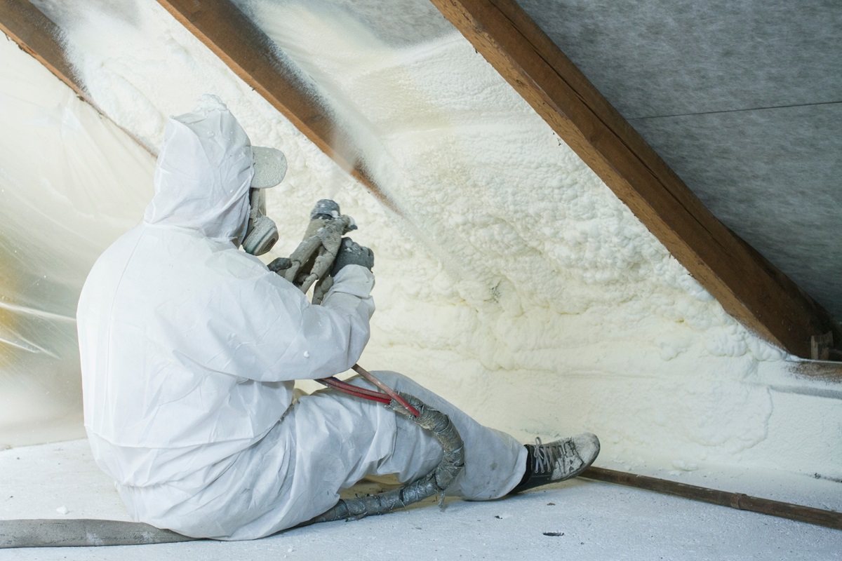 Worker sitting and using spray gun to install spray foam insulation in an attic.