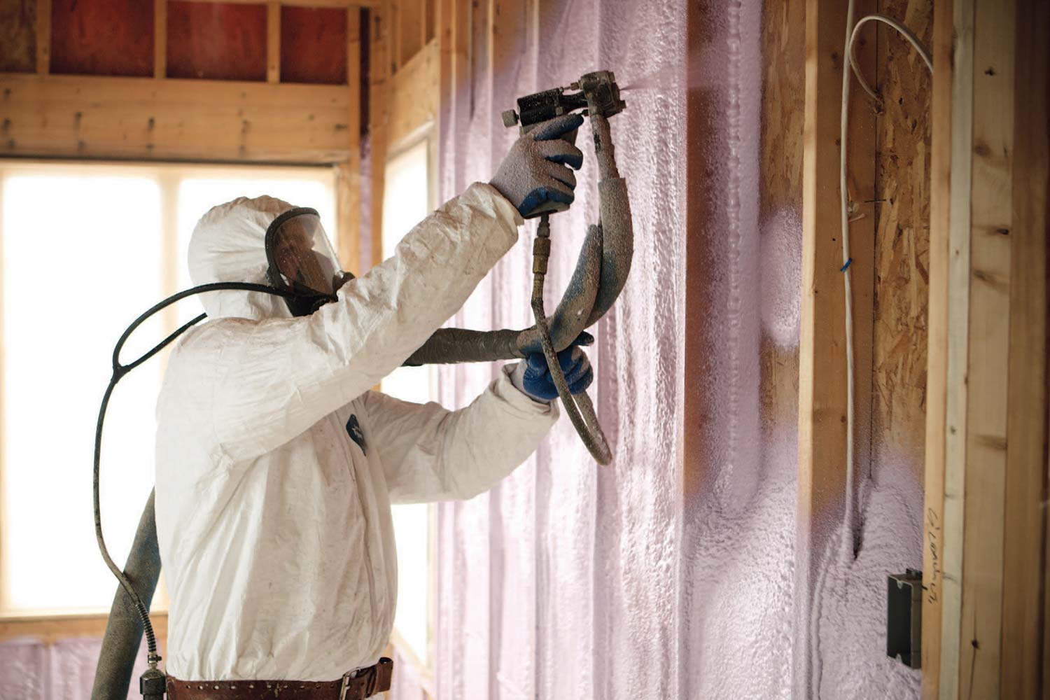 Worker using spray gun to install open cell insulation foam in residential home build.