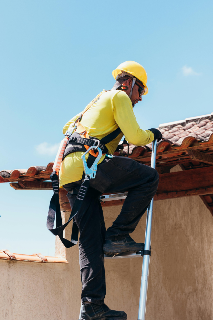 A roofing contractor in a yellow shirt and hard hat climbs a ladder onto a roof.
