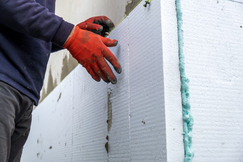 A person wearing orange gloves attaches a foam insulation board to the side of a building.