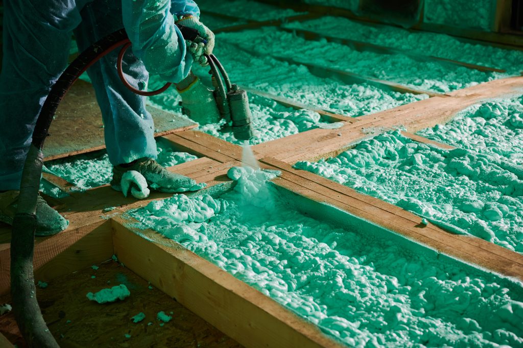 A person wearing PPE uses a machine to spray blue spray foam insulation into the frame of a building.