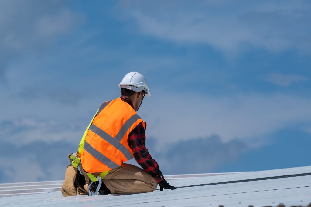 A person in an orange reflective vest and white hard hat sits on top of a white roof.