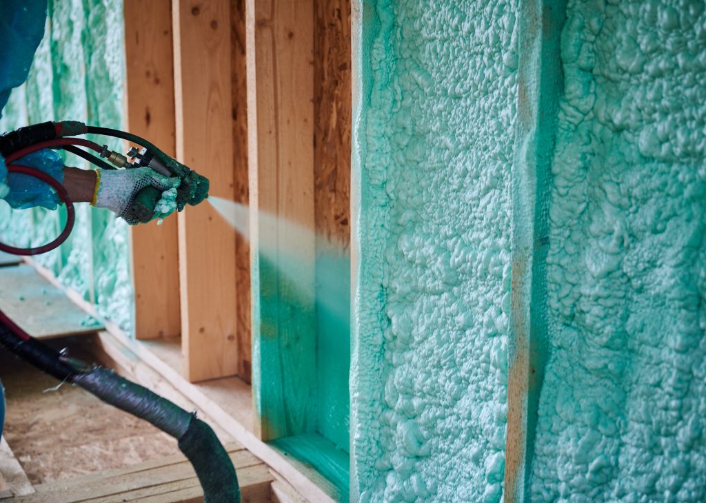 A hand wearing a work glove uses a machine to spray blue foam insulation onto a wall.