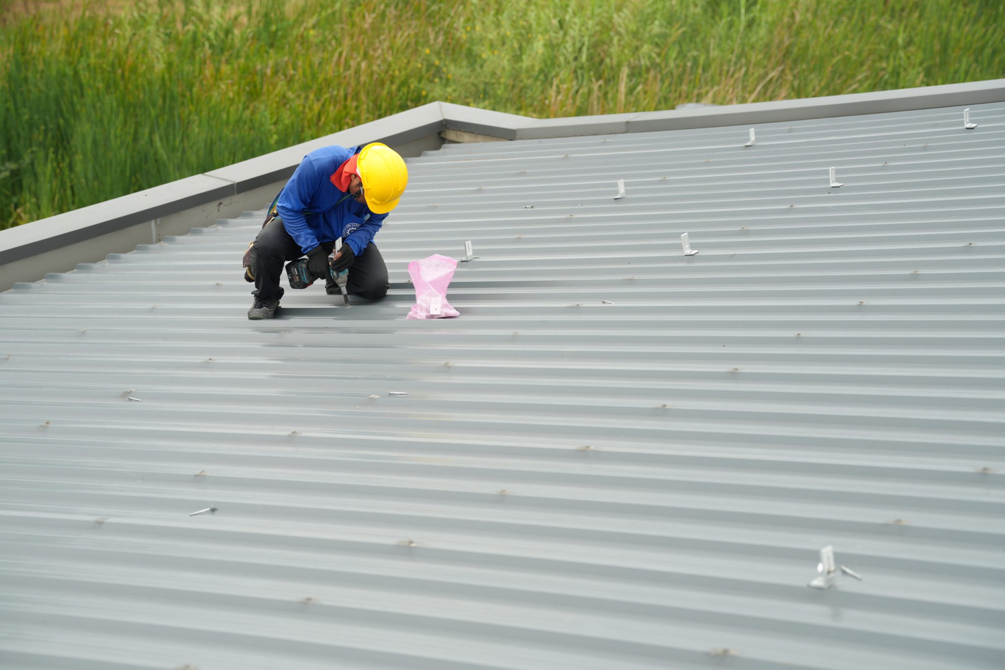 A person in a yellow hard hat uses a drill to work on a silver metal roof.