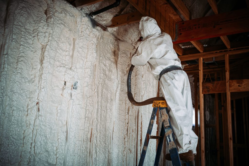 A person in white PPE adds white spray foam insulation to the walls of a building.