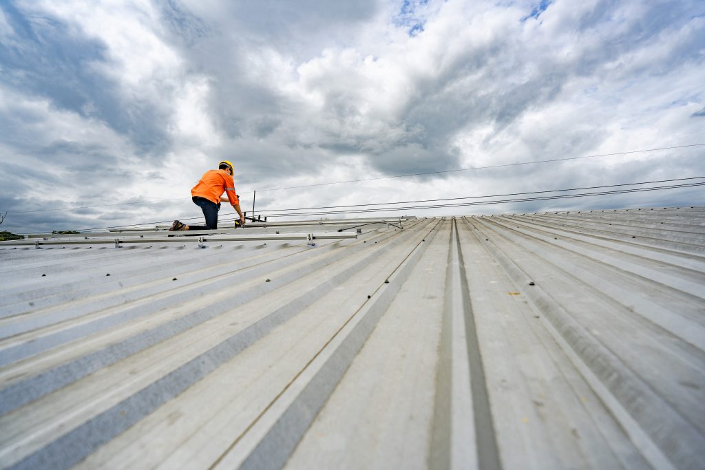 A person wearing an orange shirt and yellow hard hat works on a metal roof on a partly cloudy day.