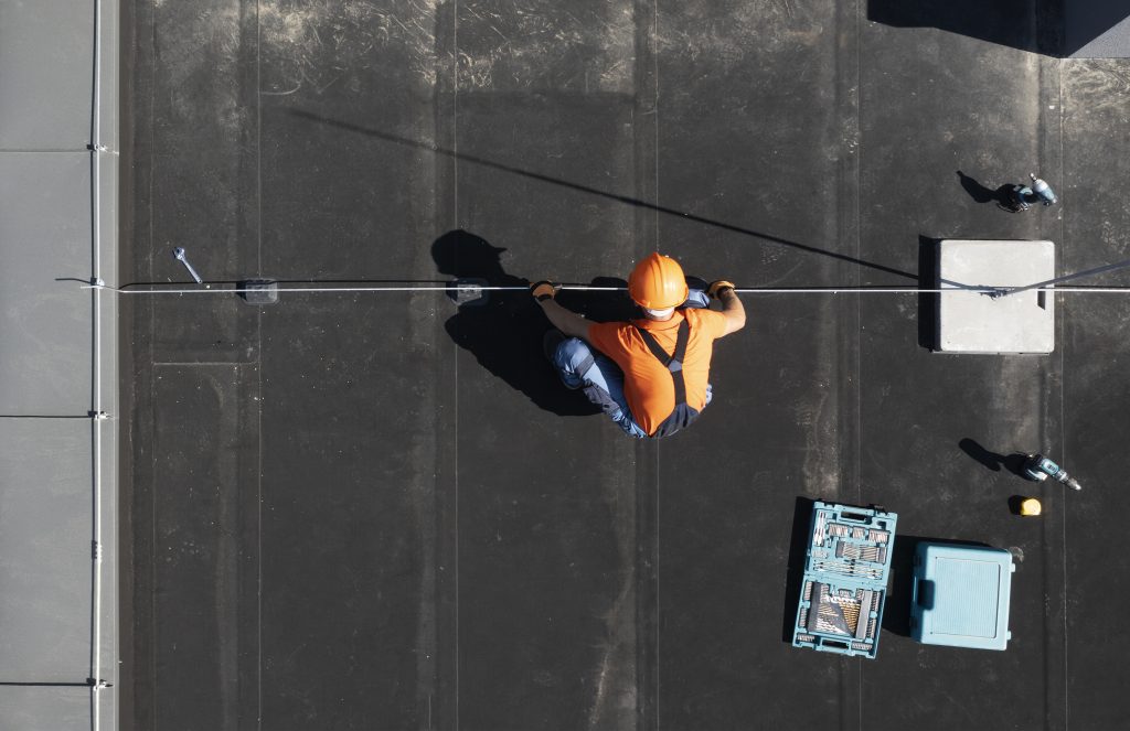 A roofing contractor in an orange hard hat works on a roof next to a blue toolbox.
